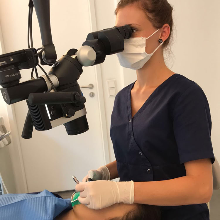 Eine Person in blauer medizinischer Kleidung und Maske arbeitet mit einem Mikroskop über einer liegenden Person.