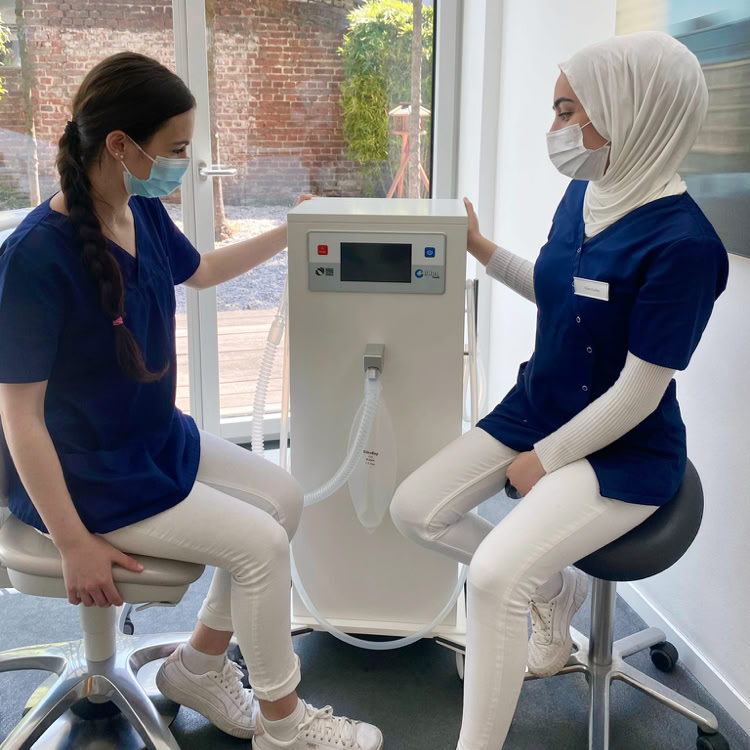 Zwei Personen in blauer Arbeitskleidung und Masken sitzen auf Hockern neben einem medizinischen Gerät in einem Raum mit großen Fenstern.